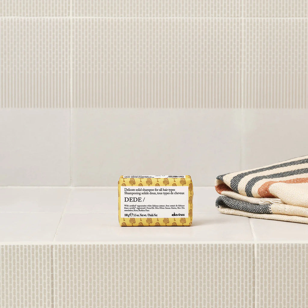 Dede Delicate Solid Shampoo Bar placerad på vitt kakel bredvid vikta handdukar med ränder, en miljövänlig fast schampoprodukt för skonsam hårvård.