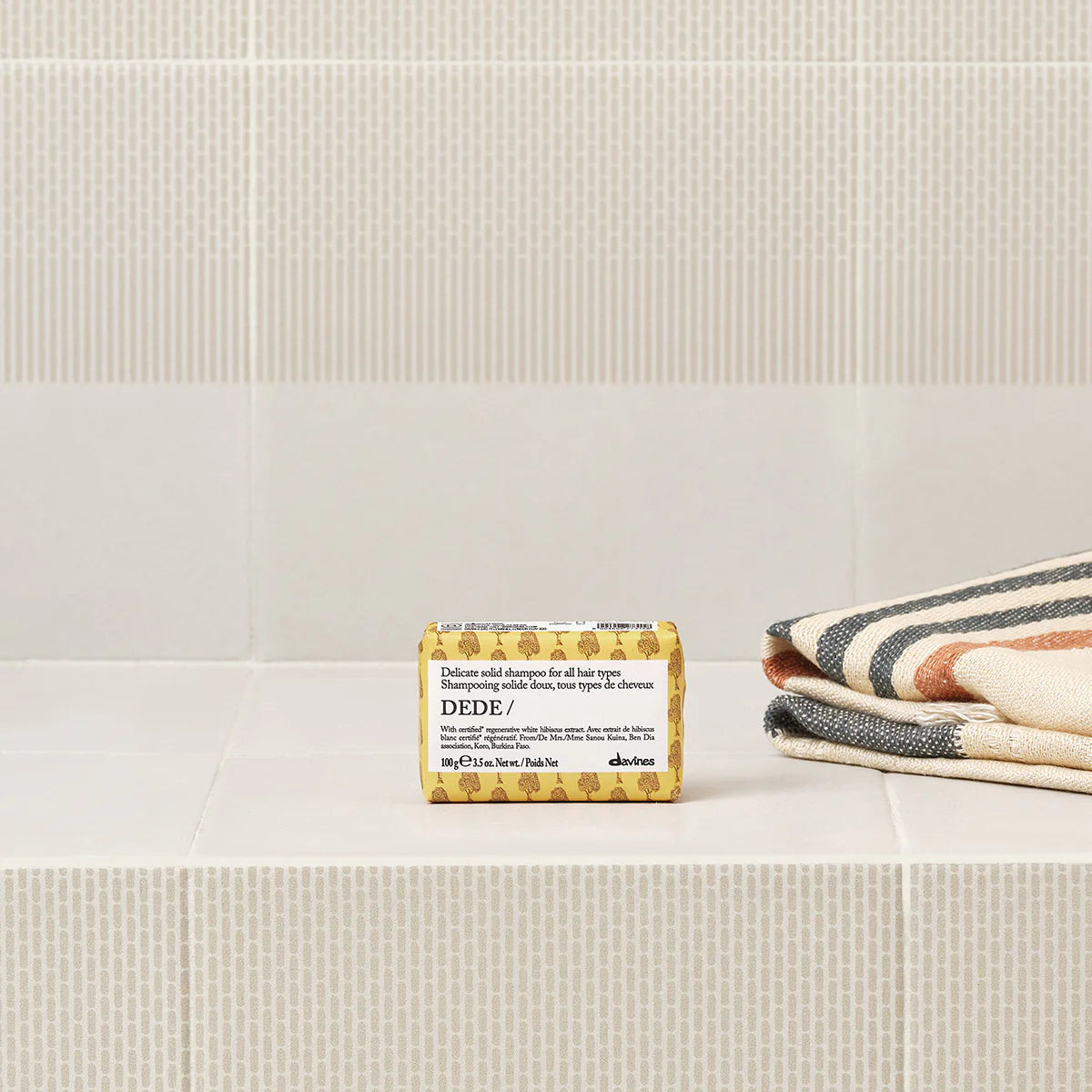 Dede Delicate Solid Shampoo Bar placerad på vitt kakel bredvid vikta handdukar med ränder, en miljövänlig fast schampoprodukt för skonsam hårvård.