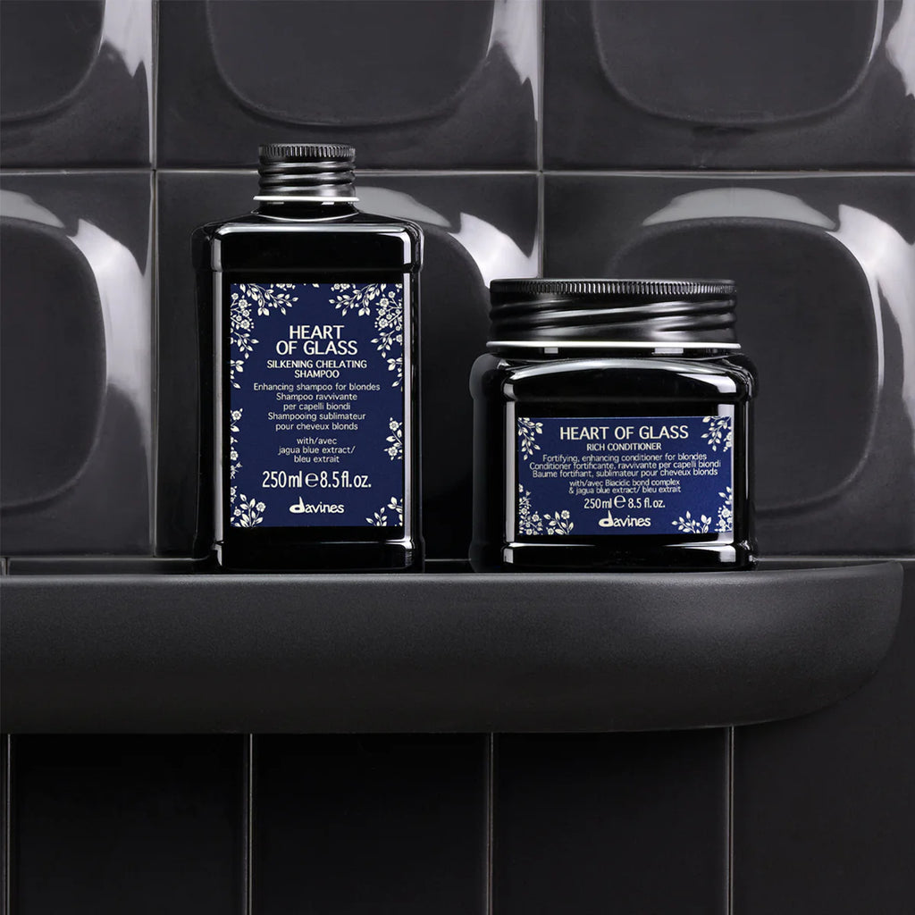 Heart of Glass Rich Conditioner bredvid en matchande hårvårdsprodukt i svarta förpackningar med elegant blå etikett, placerade på en svart hylla mot mörka kakelväggar.