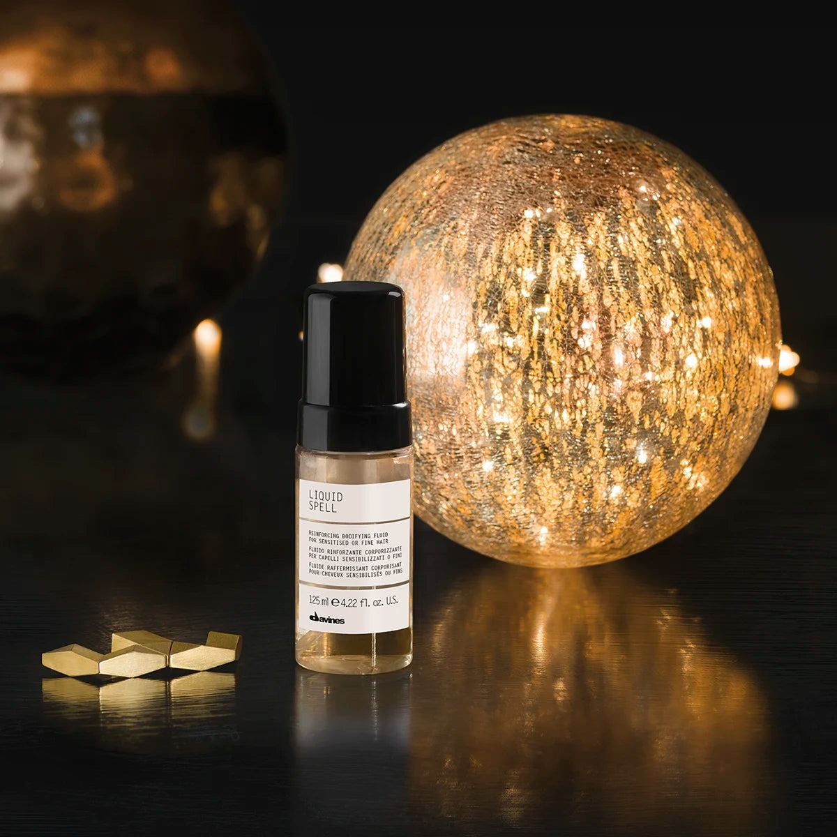 Flaska med Liquid Spell stylingprodukt med lätt textur bredvid en dekorativ, gyllene ljusboll på en mörk yta.