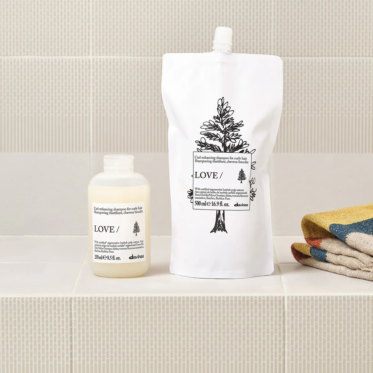 Love Curl Enhancing Shampoo i miljövänlig förpackning tillsammans med refill på ett badrumsgolv bredvid vikta handdukar, schampo för lockigt hår.