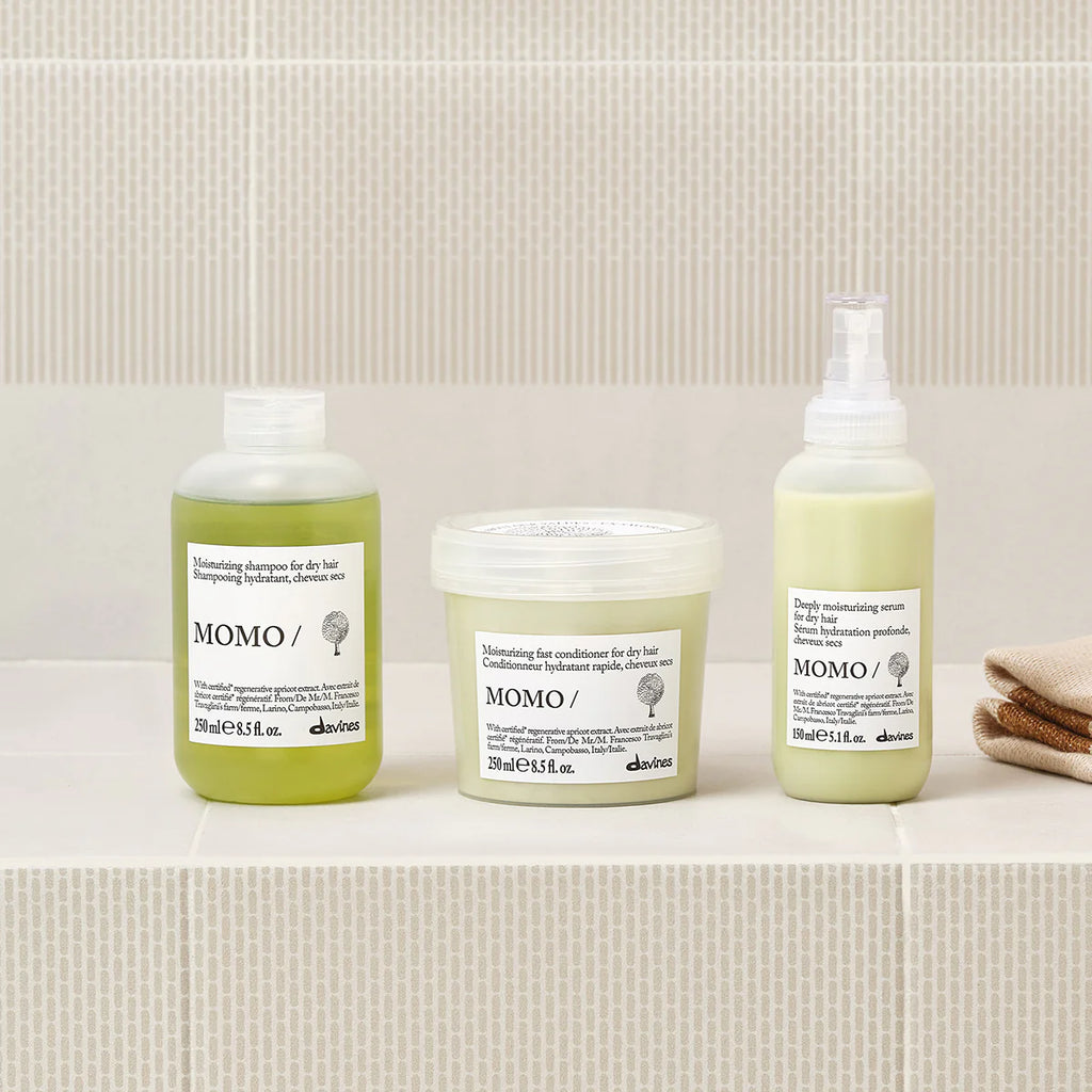 Flaskor med hårvårdsprodukter inklusive Momo Moisturizing Shampoo för torrt hår, placerade på ljus marmorhylla med neutral bakgrund och handduk.