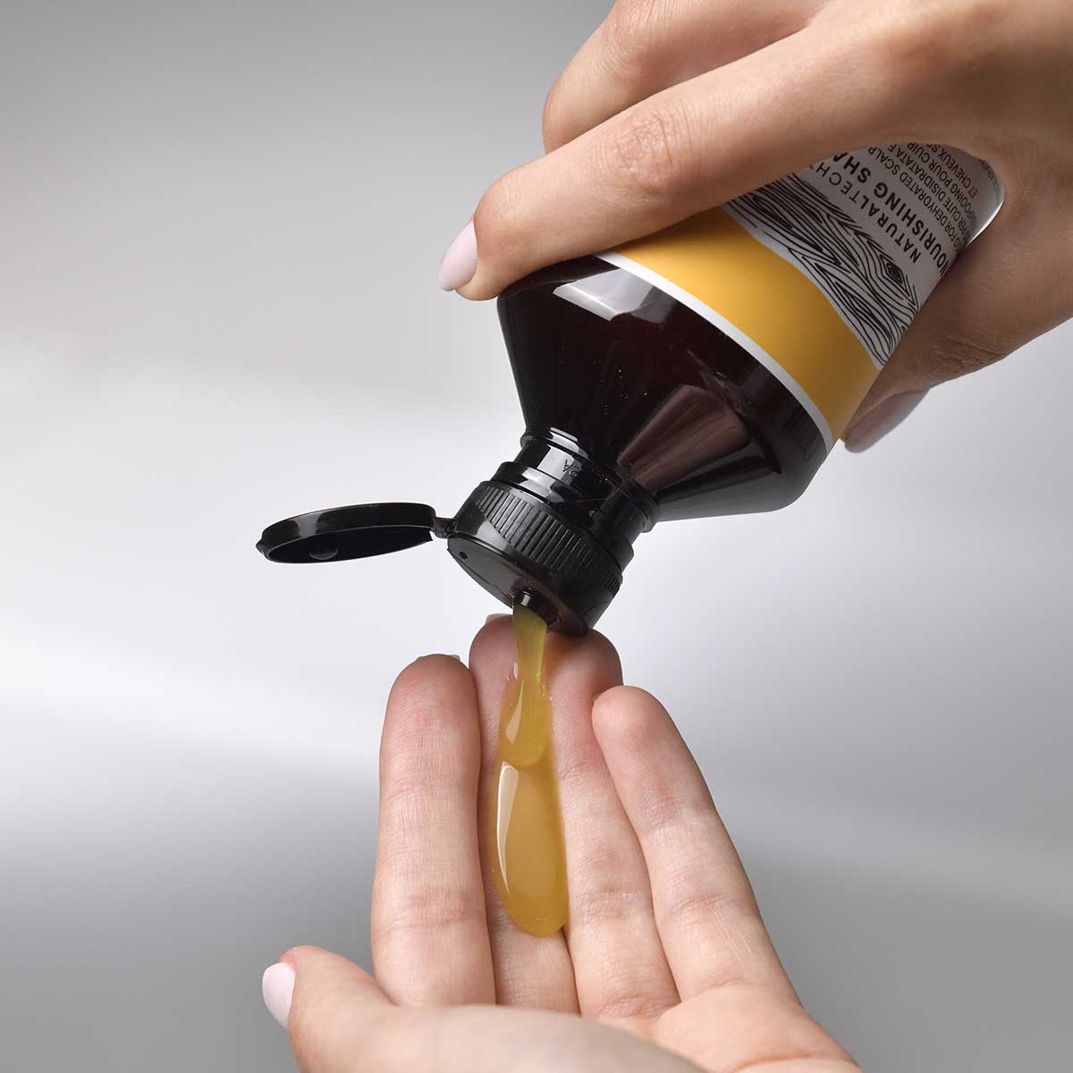En närbild på en hand som trycker ut Naturaltech Nourishing Shampoo i genomskinlig, gyllene gelé från en flaska med svart lock mot en ljus bakgrund.