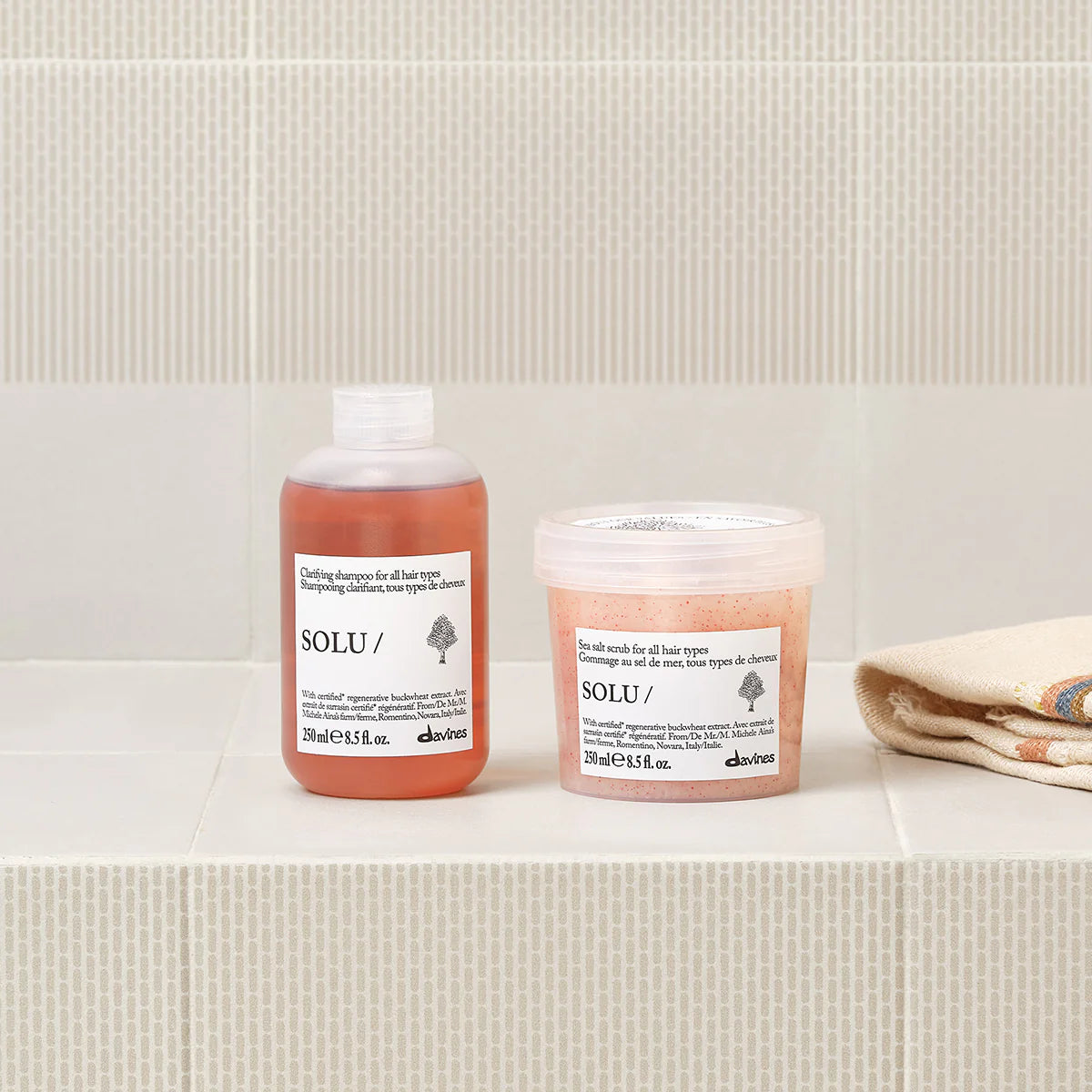 Solu Clarifying Shampoo i flaska bredvid en burk peeling för hårbotten, placerade på beige kakel tillsammans med en handduk, miljövänliga hårvårdsprodukter för rengöring och vård.