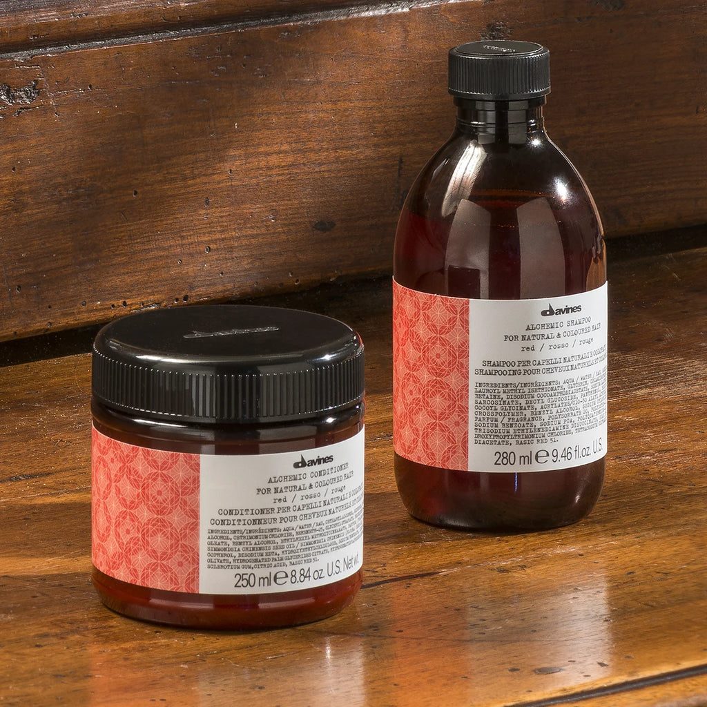 Alchemic Conditioner Red i en brun plastburk med svart lock, placerad på träbord bredvid matchande schampoflaska med etikett i rött och vitt för färgat hår.