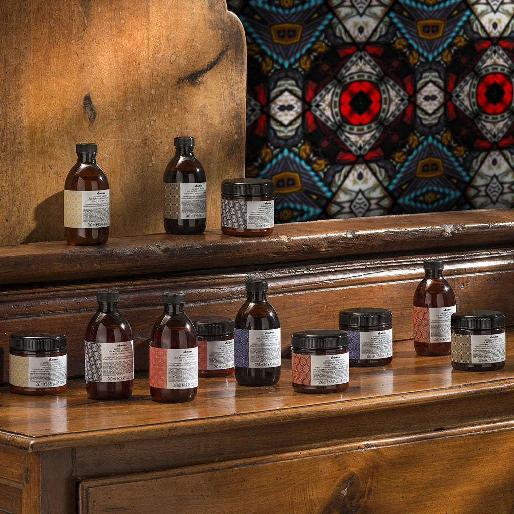 Alchemic Shampoo Silver tillsammans med andra hårvårdsprodukter från Davines i glasflaskor och burkar placerade på ett träbord med färgstarkt bakgrundstyg.